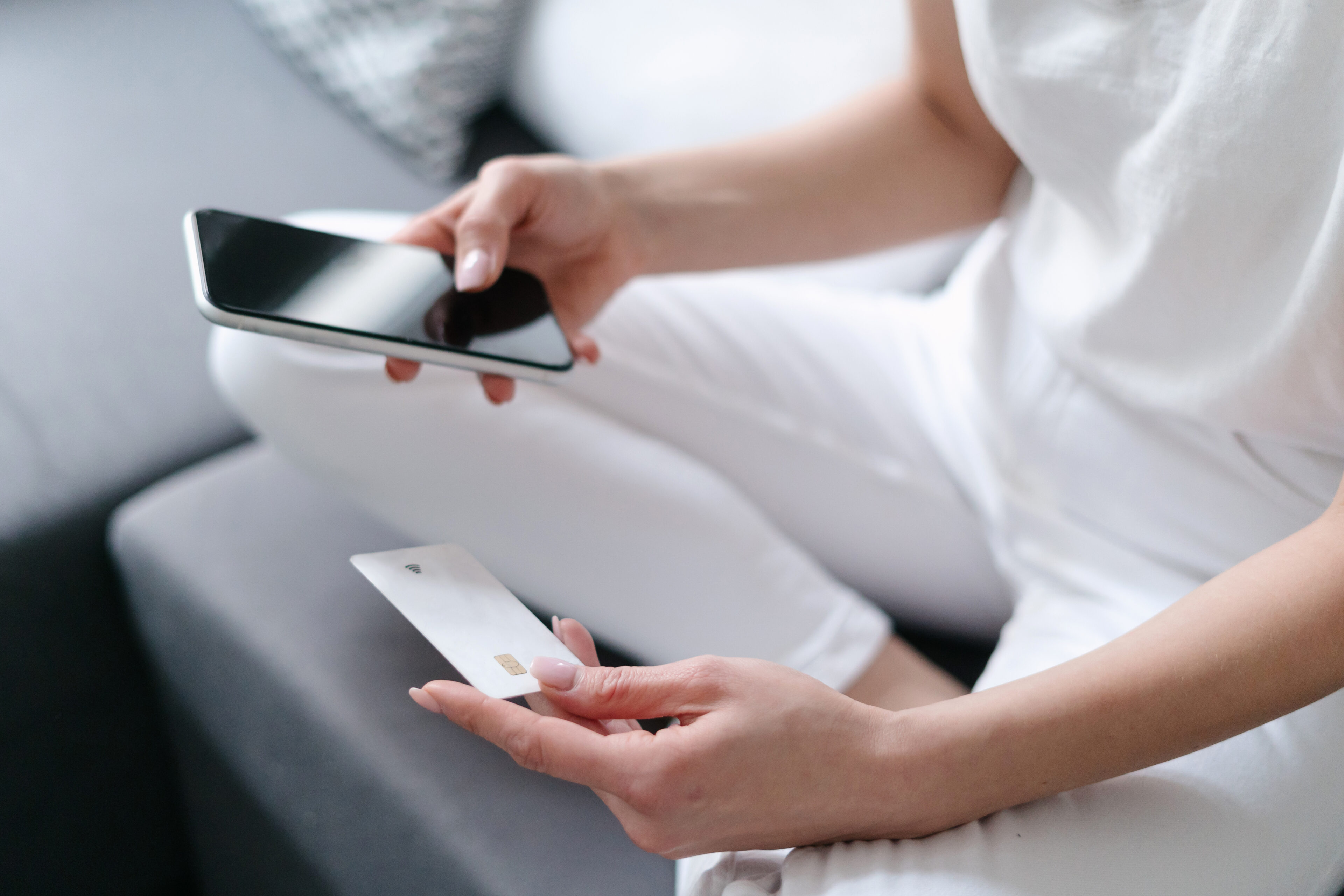 cropped shot of female hand holding credit card and using smart phone for online purchase while sitting on sofa at home, money transfer, donate and charity concept