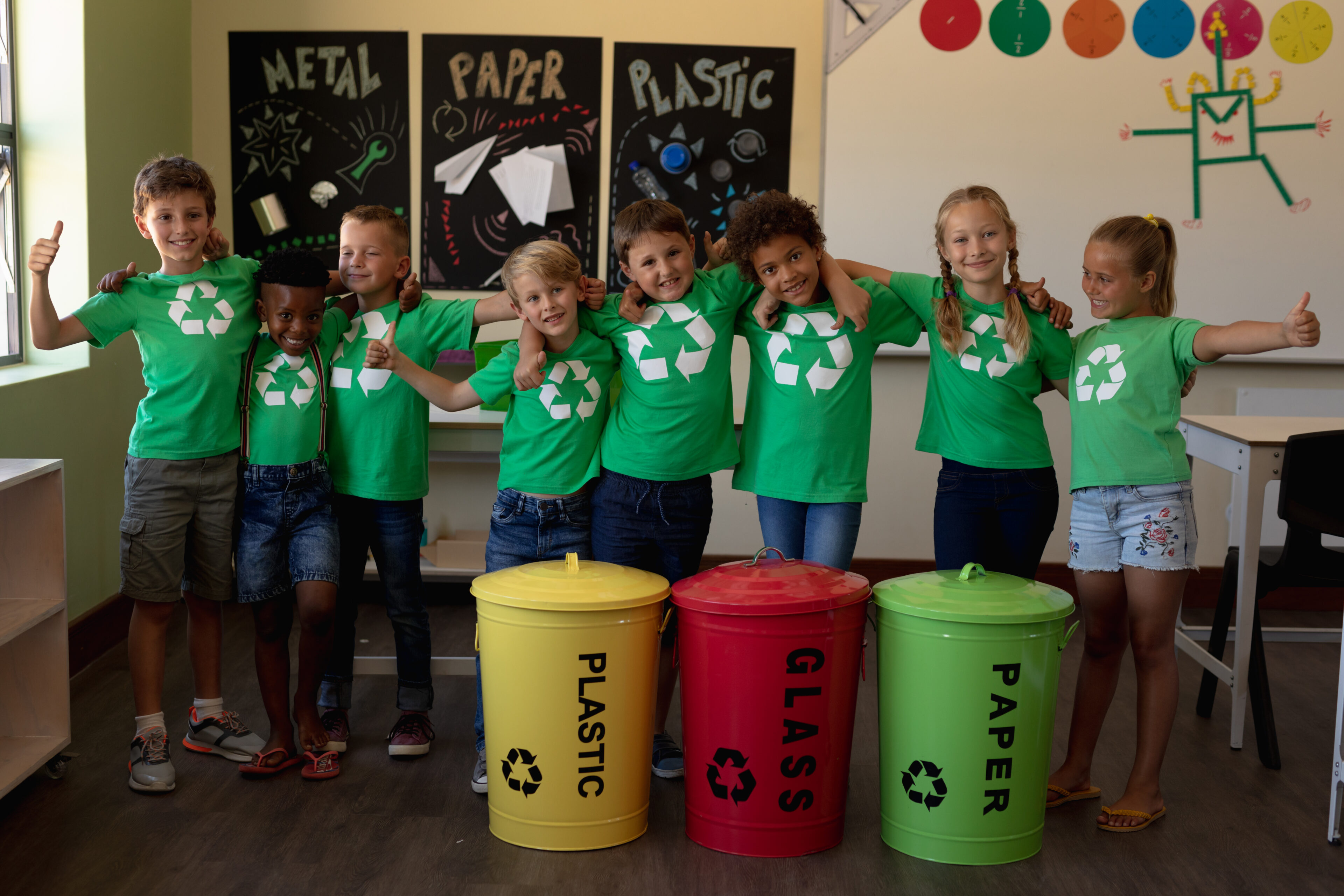 Portrait of a diverse group of schoolchildren wearing green t shirts with a white recycling logo on them, standing behind colour coded recycling bins in an elementary school classroom with arms around each other, smiling to camera with thumbs up