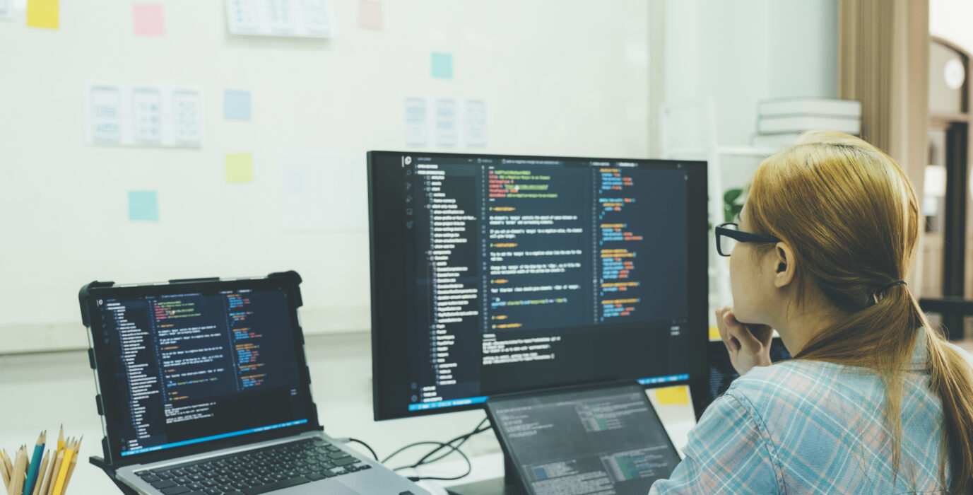 Female developer sits coding at multiple screens.